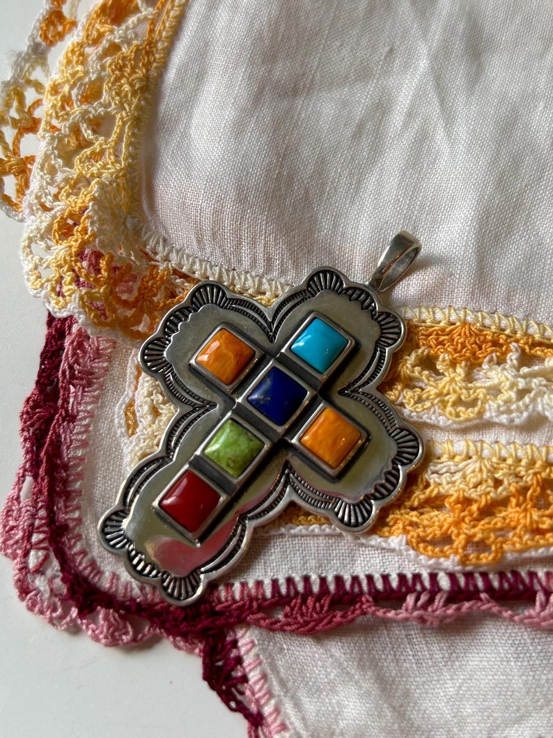 May include: A silver cross pendant with a southwestern design. The cross is decorated with eight colorful stones in a variety of colors, including red, orange, yellow, green, blue, and turquoise.