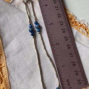 May include: A silver necklace with blue beads. The necklace is approximately 5 inches long. The beads are a deep blue color and are interspersed with silver tube-shaped links. The necklace is displayed on a white cloth with a yellow lace trim.