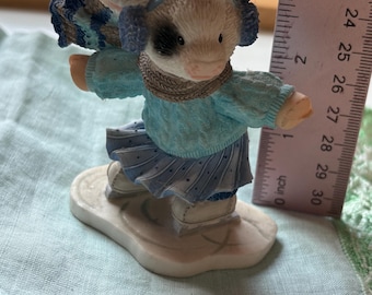 May include: A figurine of a cow wearing a blue sweater, scarf, earmuffs, and skirt, standing on a white base. The cow has black and white markings. The figurine is approximately 2 inches tall.