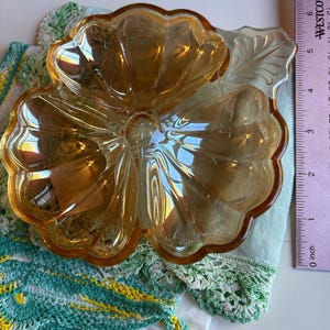 May include: A clear, amber-colored glass serving dish in the shape of a flower with three petals and a leaf. The dish is resting on a green and white crocheted doily. A ruler is next to the dish.