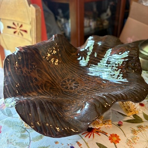 May include: A dark brown ceramic bowl with a glossy finish and a wavy, organic rim. The interior surface features a textured wood grain pattern and a small, stamped floral design at the center, creating a deep, bowl-shaped vessel.