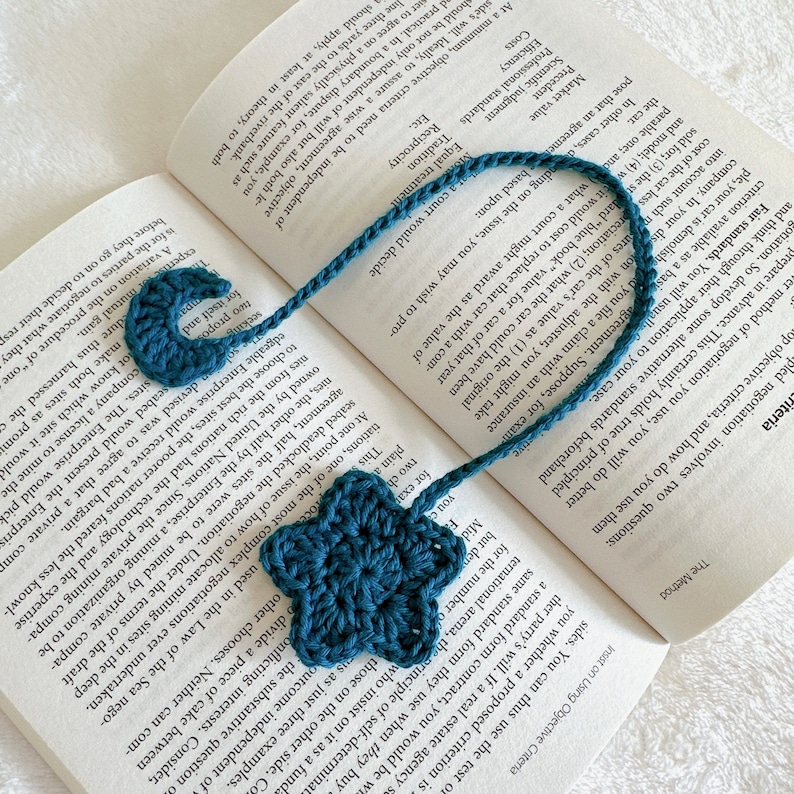 Puede incluir: Un marcap&aacute;ginas de color azul verdoso tejido a crochet con un dise&ntilde;o de estrella y media luna. El marcap&aacute;ginas est&aacute; hecho de hilo y tiene un cord&oacute;n largo para sujetarlo a un libro.