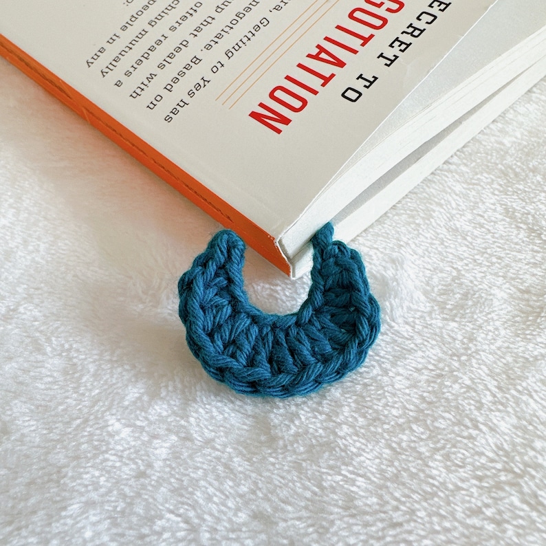 Puede incluir: Un marcap&aacute;ginas de media luna de color azul turquesa tejido a crochet se coloca sobre una superficie blanca y peluda. El marcap&aacute;ginas est&aacute; apoyado sobre un libro cerrado con el t&iacute;tulo "Secret to Negotiation" visible.