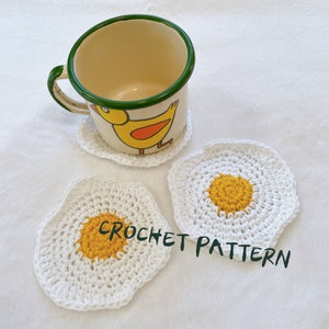 Puede incluir: Dos posavasos de crochet blanco con forma de huevos fritos con centros de hilo amarillo. Una taza de esmalte vintage con un diseño de pollito amarillo se encuentra sobre un posavasos de crochet blanco.