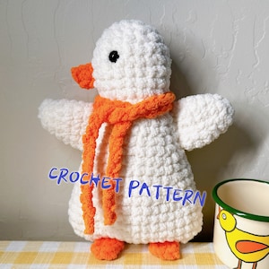 May include: White crocheted penguin amigurumi toy with an orange scarf. The penguin has black eyes and an orange beak. The toy is sitting on a yellow and white checkered surface. CROCHET PATTERN is written in blue on the image.
