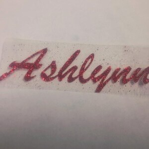 May include: A red glitter iron-on transfer decal featuring the name Ashlynn written in a cursive script font. The letters are textured with fine red sparkles and are set against a clear, rectangular adhesive backing material.
