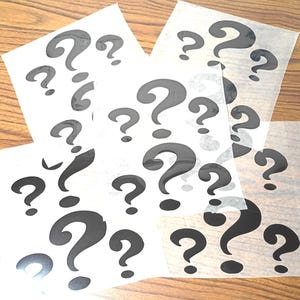 May include: Several sheets of white paper are scattered on a wooden surface, each printed with a repeating pattern of black question marks. The question marks vary in size and are arranged in a random, overlapping manner.