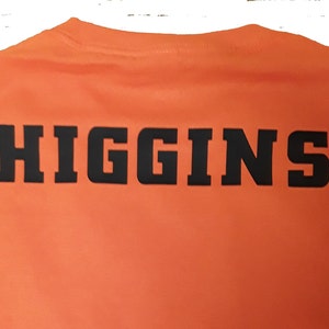 May include: Orange t-shirt with the word "HIGGINS" printed in black on the back.