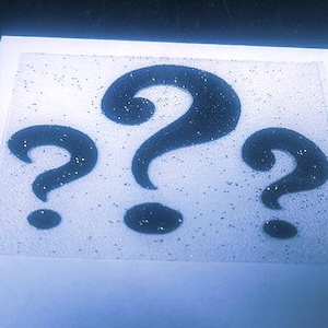 May include: A rectangular, textured, semi-transparent sheet featuring three dark blue question marks arranged horizontally. The surface of the sheet is covered in a fine, reflective glitter finish that sparkles against the light blue background.