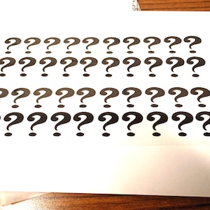 May include: A white sheet of paper with rows of black question marks printed on it. The question marks are evenly spaced and arranged in a grid pattern. The paper is on a wooden surface.