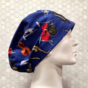 May include: A blue scrub cap featuring a repeating pattern of musical instruments including a red drum set with the text The Jazz Lounge, a brown cello, a saxophone, a piano, and a trumpet. The cap is displayed on a white mannequin head.