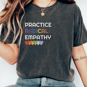May include: A dark gray t-shirt with the text "PRACTICE RADICAL EMPATHY" in white letters. The text is surrounded by a row of rainbow-colored hearts.