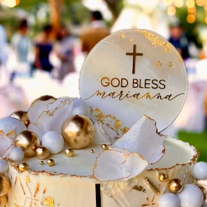 May include: A white cake decorated with gold accents, white and gold spheres, and a white sign that says "God Bless Marianna" with a gold cross on top.