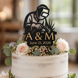 May include: Wedding cake with a decorative topper featuring a silhouette of a welder and the initials "A & M" with the date "June 21, 2026". The cake is decorated with white frosting, roses, and greenery.