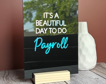 May include: A black acrylic sign with the text "IT'S A BEAUTIFUL DAY TO DO Payroll" in white and teal lettering. The sign is propped up by a small wooden stand. A white vase is to the right of the sign.