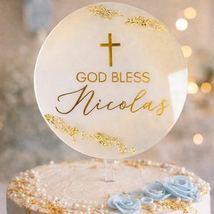 May include: A round cake topper with the words "GOD BLESS Nicolas" in gold script, topped with a gold cross and gold leaf accents. The cake is white with blue frosting and blue flower decorations.