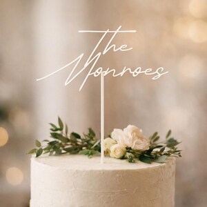 May include: A white cake topper with the words "The Monroes" in a cursive font. The topper is placed on a white frosted cake decorated with greenery and white flowers. The cake is a centerpiece for a wedding or celebration.
