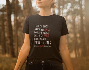 May include: A dark gray t-shirt with the text "FOOL ME ONCE SHAME ON YOU FOOL ME TWICE SHAME ON ME BUT FOOL ME THREE TIMES AND THEY'LL NEVER FIND YOUR BODY" in white and red lettering.