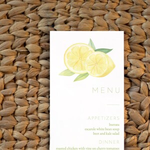 May include: A white menu card with a watercolor illustration of two lemon slices. The menu lists appetizers, dinner, and dessert options. The appetizers include burrata, escarole white bean soup, and beet and kale salad. The dinner options are roasted chicken with vine on cherry tomatoes, wood grilled salmon with sauteed spinach, rigatoni with vodka sauce, and cacio e pepe. The dessert options are funfetti birthday cake and crème brûlée.