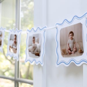 May include: A photo banner featuring multiple square cards with wavy blue borders strung together on a white cord. Each card displays a photograph of a baby in a neutral-colored outfit, with one card showing a baby sitting next to a wooden block with the number 9.