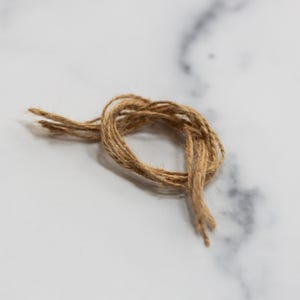 May include: Close-up of a natural brown twine tied in a knot. The twine is made of several strands and has a slightly frayed texture. The knot is centered and takes up a small portion of the frame. The background is a white and gray marble surface.