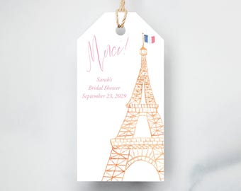 May include: A white gift tag with a watercolor illustration of the Eiffel Tower in gold. The tag includes the word "Merci!" in pink script, along with the text "Sarah's Bridal Shower September 23, 2029."