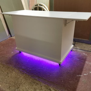 May include: White bar cart with a white top and a purple LED light strip underneath. The cart is on wheels and has a shelf.