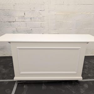 May include: A white rectangular counter or table with a flat top and a decorative panel on the front. The panel has a raised rectangular border. The table is painted in a solid, neutral white colour and is set against a white brick wall.