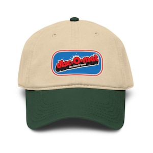May include: A beige baseball cap with a dark green brim. The cap features a rectangular patch with the words "disc-o-mat RECORDS & TAPES" in red, black, and blue. The cap has a dark green button on top.