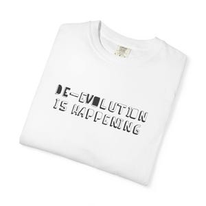 May include: White t-shirt with the text "DE-EVOLUTION IS HAPPENING" in black, block letters. The shirt is folded and appears to be made of a soft material. A tag is visible at the neckline.