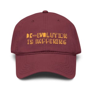 May include: A maroon baseball cap with the text "DE-EVOLUTION IS HAPPENING" in a yellow-orange font. The cap has a curved brim and a rounded crown, suitable for casual wear.