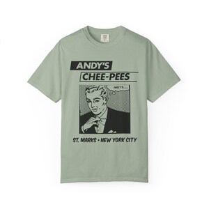 May include: A sage green short-sleeve t-shirt featuring a black graphic print. The design includes the text ANDY'S CHEE-PEES, a comic-style illustration of a person in a suit, a thought bubble with the text ANDY'S, and ST. MARKS • NEW YORK CITY.