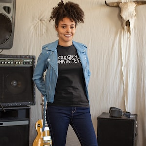 May include: A woman wearing a black t-shirt with the text "OLD GREY WHISTLE TEST" in white lettering. She is wearing a light blue denim jacket and blue jeans. The woman is standing in front of a wall with a bull skull hanging on it.