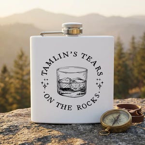 Könnte beinhalten: Eine weiße Flasche mit silbernem Verschluss und Scharnier, mit dem Text "TAMLIN'S TEARS ON THE ROCKS" um eine Grafik eines Whiskyglases. Ein antiker Kompass und ein Lederriemen liegen neben der Flasche.