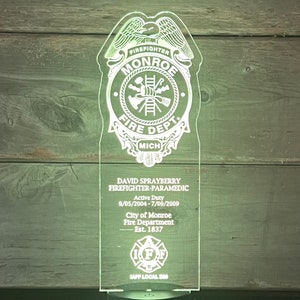 Puede incluir: Una placa de acrílico blanco con una base negra, que presenta una insignia de bombero con las palabras "Monroe Fire Dept. Mich." y el nombre de un bombero, las fechas de servicio y "IAFF Local 300".