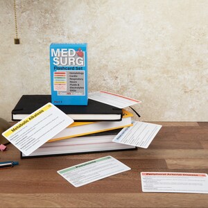 May include: A blue box of "MED SURG Flashcard Set" with flashcards scattered on a wooden surface. The flashcards are labeled with medical terms like "Metabolic Alkalosis" and "Pneumonia". A desk lamp and books are also visible.