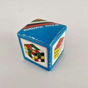 May include: A blue box with the words "WONDERFUL PUZZLER" printed on it. The box features illustrations of a Rubik's Cube on its sides. The cube is a classic puzzle with coloured squares. The box is cube-shaped.