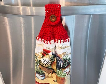 Op de afbeelding: Rode gehaakte theedoek met een knoop en een haanontwerp. De handdoek zegt "Rooster" en heeft een rode rand.