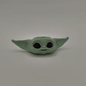 May include: A green 3D printed figurine of a character from the Star Wars franchise, known as "The Child" or "Baby Yoda". The figurine has large eyes and a small mouth, and is in a sitting position.