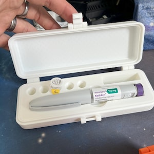 Può includere: Un astuccio rettangolare bianco aperto, contenente una penna per iniezione preriempita e una piccola fiala. La penna è grigia con un cappuccio viola e un'etichetta verde che recita "mounjaro 7,5 mg KwikPen". La fiala è gialla e bianca.