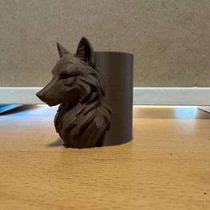 Könnte beinhalten: Ein brauner, zylindrischer Behälter mit einer detaillierten Wolfskopfskulptur auf der Vorderseite. Die Merkmale des Wolfes sind aufwendig geschnitzt und zeigen sein Fell und seine Gesichtsstruktur. Der Behälter ist wahrscheinlich ein dekorativer Gegenstand oder ein kleiner Pflanztopf.