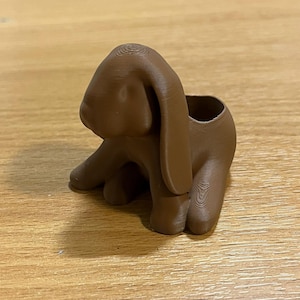 Puede incluir: Un macetero de conejo marrón impreso en 3D. El conejo está en posición sentada con orejas largas y caídas y una pequeña abertura en la espalda para plantar. El macetero es de material liso.