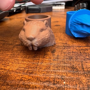 Könnte beinhalten: Ein brauner 3D-gedruckter Ring mit einem geformten Biberkopf, sichtbarer Schichtstruktur und zwei markanten Vorderzähnen. Daneben befindet sich ein blauer 3D-gedruckter Ring mit einem geformten Adlerkopf. Beide Ringe haben ein zylindrisches Banddesign.