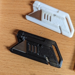 May include: Two 3D printed utility knives, one black and one white, with a silver blade. The knives have a textured surface and a small hole for attaching a lanyard.