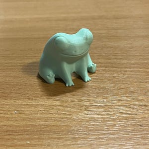 May include: A light green frog figurine with a smiling expression. The frog is sitting upright with its arms and legs extended. The figurine is made of a solid material and has a textured surface.