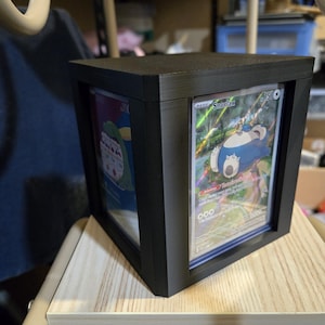 May include: A black, cube-shaped display case holding two Pokémon trading cards. One card features a blue and white Snorlax character. The case is made of a matte black material and is designed to showcase the cards.