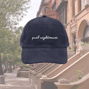 Puede incluir: Gorra de béisbol de pana azul marino con la frase "quel nightmare" bordada en blanco. La gorra tiene una visera curva y una corona estructurada. Un accesorio elegante para el uso diario.