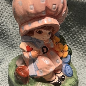 May include: A ceramic figurine of a girl wearing a pink and white polka dot dress and a large pink hat with white polka dots. She is holding a red heart and a blue and white letter. She is sitting on a green base with a brown log and a blue watering can.