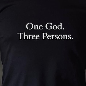 Puede incluir: Camiseta negra con el texto blanco "One God. Three Persons." La camiseta es de manga corta y parece estar hecha de un material suave. La persona lleva pantalones color caqui y un reloj negro.