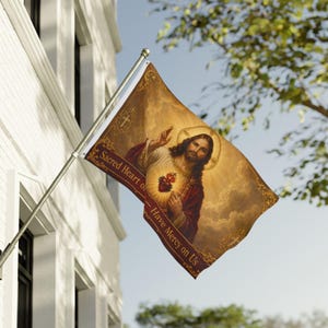 May include: A flag featuring a religious image of Jesus Christ with a halo and a heart. The flag has a brown background with the text "Sacred Heart of - Have Mercy on Us". The flag is mounted on a silver pole and is displayed outdoors.